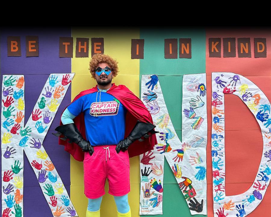 A handmade sign that reads "Be the I in kind". Captain Kindness stands in front of the sign.